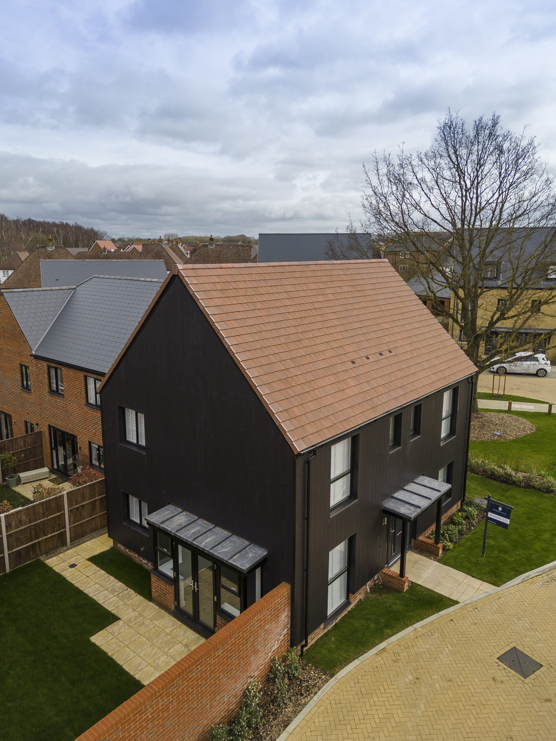 Aerial view of a modern, dark-colored house with a light red sloped roof, surrounded by a neatly landscaped yard with green grass and paved walkways. Nearby houses with similar architectural styles are visible in the background under a cloudy sky